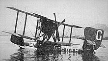 Black-and-white photograph of a biplane, facing away from the camera, floating on calm water.