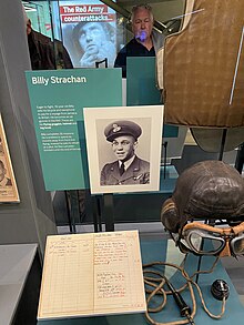 A photograph of a museum display containing items once belonging to Billy Strachan. On this display there are flight goggles and a flight helmet, a log book, and a photograph of Billy Strachan