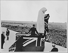 In the black and white photograph, the unveiling of the Vimy Memorial is captured. A figure stands atop a stage draped in flags, positioned in front of the poignant statue of Canada Bereft. A vast crowd of thousands of people are gathered and are facing towards the stage.