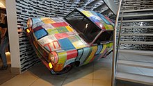 A car with bright coloured squares painted on the exterior is tilted slightly to its left side at the bottom of a spiral staircase.
