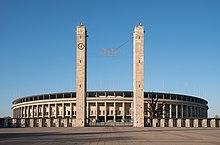 Frontale Farbfotografie vom Olympiastadion mit seinem hellen Gebäude und Stützsäulen. Vor dem runden Sandsteingebäude sind in dem Zaun zwei schmale Türme integriert, die durch Drähte die Olympischen Ringe in der Mitte festhalten.