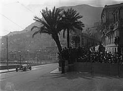 A large, open sports car drives past crowds of people and palm trees, with the city of Monaco in the background