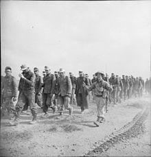 A column of German prisoners walk, from right to left, escorted by a British soldier giving two thumbs up.