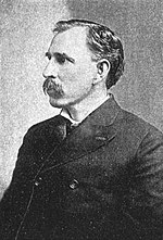 A mustachioed man in his mid-forties with black hair. He is wearing a black coat buttoned at the high chest and facing left