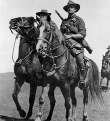 Black and white photo of a man wearing military uniform with a rifle slung across his back riding a horse. Two other similarly dressed men are partly visible in the background.