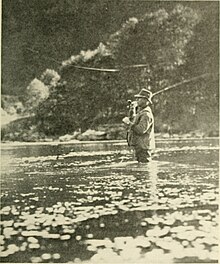 A man wearing waders, standing thigh deep in the river casting a flyrod