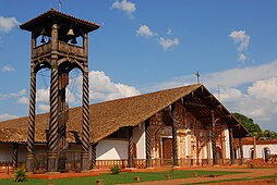 A wooden bell tower and a church in three-quarter view. The bell tower consists of a roof-covered platform supported by four columns with twisted fluting. Clocks are attached to the platform and a spiral staircase leads to it. The facade of the church is white and decorated with orange paintings. The church roof is rather large.