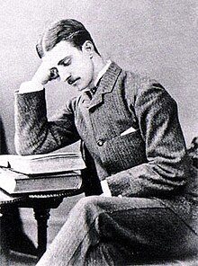 Druitt, a thin young man with dark hair parted in the middle and a small moustache, sits at a table and reads a book