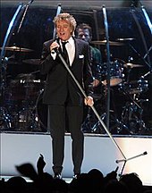 A man on a stage, wearing a suit and holding a mic and its stand at an angle. Behind him, a man is playing a drum set.