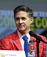 A middle aged caucasian male looks slightly to the leftwith a small smile. He has short brown hair and is wearing a Spider-Man-themed red coat with webbing trim.
