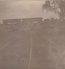 A small steam locomotive and tender, along with two passenger cars, parked in front of an open field. Stairs connect the field to the railroad tracks, which are at a higher elevation.
