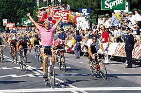 Fignon, wearing a pink jersey, throwing his arms up in celebration as he crosses the line in front of other riders.