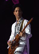 Hip height portrait of Prince playing guitar at night wearing white suit with metallic silver ornament