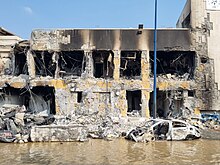 A severely-damaged building, looking as if it were a cross-section. In front is a burned car partially submerged in murky brown water.