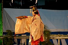 Sur une scène de Takigi nō (scène de plein air à structure de bambou) l'acteur porte un kimono orange revêtu d'un surplis jaune pâle, dont les manches amples cachent sa main, qui agite un éventail. Il porte un masque traditionnel et un couvre-chef doré, surmonté d'un papillon et orné de breloques clinquantes