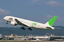 A Boeing 777-200 aircraft in mid-air during take-off, with the view of Itami Airport in the background