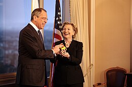 Hillary Clinton standing with Russian Foreign Minister Serguéi Lavrov. Both of them are holding a "reset button". They are in a room with a window to the left and an American flag behind them