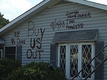 Damaged white home, with "Buy Us Out" and "We R Done" written on the siding