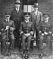 Formal portrait of five men, three seated and wearing military uniforms with peaked caps, two standing and wearing civilian clothes and hats