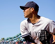 Mariano Rivera in a gray baseball uniform, navy blue cap, and baseball glove bearing his name. He is finishing his throwing motion to the left, and is squinting in the daytime sunny conditions.