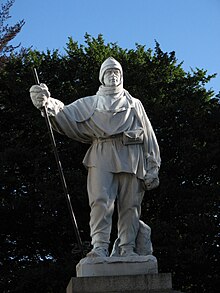 A white statue of Antarctic explorer Robert Falcon Scott stands in front of a group of trees. The statue is white and shows Scott wearing many layers of fur and other clothing, as he would have done while in Antarctica.