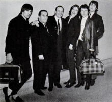 The Kinks and the two executives, all in suits, pose for a group photo, luggage in hand.