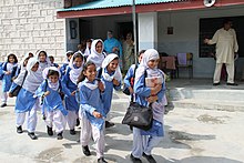 Pakistanske jenter i skuleuniform som består av blå kamiz og kvite salvarbukser og dupatta. Mannen i bakgrunnen ber heilkvit salvar kamiz. Foto: Vicki Francis/Department for International Development