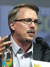 A man with dark brown hair, sporting rectangular glasses, a mustache, and a goatee while wearing a teal shirt and dark grey glasses, looks to the right while speaking into a microphone.