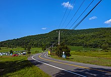 A two-lane paved road winding through countryside from just right of the camera, down the center of the frame, towards a hill covered with green trees under a blue sky with some small clouds in it. On the far side of the road there is a sign with the number 22 on it; below it is a white on blue sign with "Be Prepared to Stop" on it in capital letters. Telephone wires enter the image from top left, connecting to a wooden pole at the center