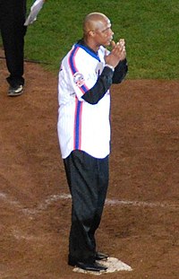 A man in black pants and a white baseball uniform with blue and orange stripes down the side holds his hands close to his face while standing on home plate.