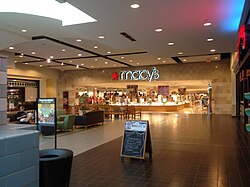 A one-story shopping mall concourse with brown tile flooring. Present is the entrance to a Macy's department store, with signage reading "Macy's"