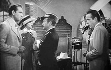 Black-and-white film screenshot of several people in a nightclub. A man on the far left is wearing a suit and has a woman standing next to him wearing a hat and dress. A man at the center is looking at the man on the left. A man on the far right is wearing a suit and looking at the other people.