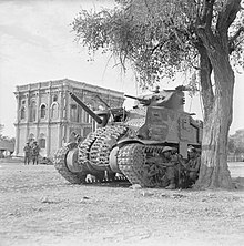 photographie en noir et blanc montrant un char à côté d’un arbre avec un bâtiment à l’arrière-plan