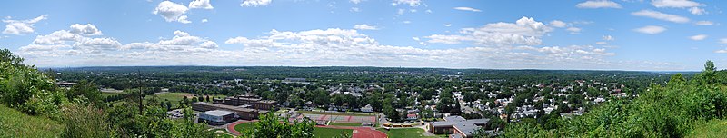 A wide panoramic shot begins and ends in bushes but is graced by two and three story city houses throughout most of the image. Front and center are three large schools featuring a track, tennis courts, and a football field. Behind the houses is a river flowing right to left.
