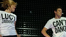 Agron and Cory Monteith, as their characters from Glee, dance enthusiastically wearing simple white T-shirts with black text: Agron's reads "Lucy Caboosey" and Monteith's "Can't Dance".