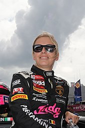A woman in her late twenties wearing black racing overalls with sponsors logo smiling to her right and wearing black sunglasses