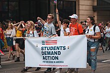 Five young adults march holding a banner that reads "Students Demand Action for Gun Sense in America". In the background, other people march and look on as the parade they are marching in passes by.