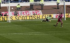 A football player scores a goal against the opposing goalkeeper from a penalty-kick. Stewards and camera-operators are visible behind the goal net.