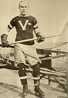 A man poses for a photo wearing skates and an ice hockey sweater, and holding a stick in his hands