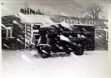 A black and white photo of a truck with a rounded front sitting in the snow in front of a one-story brick building with the words "CANNING SETS" on its outer wall