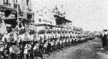 Photographie d'une colonne de soldats noirs avec leurs fusils à l'épaule et portant des chemises, des shorts et des calots clairs dans une rue bordée de bâtiments de style coloniaux