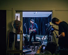 Peter plays a guitar in a room, another man is adjusting a camera on a tripod stand