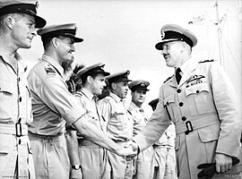 Two men in light-coloured military uniforms with peaked caps, shaking hands in front of a row of similarly dressed men