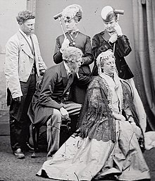 Five people in stage costumes; three are standing behind two who are seated