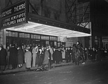 Black and white photo of a crowd of people standing on the sidewalk in front of a building that has a lighted marquee saying 'Biltmore Theatre'.