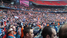 A three-tiered football stadium stand, the bottom two full of people clad mostly in white and orange. Several white and orange flags are visible.