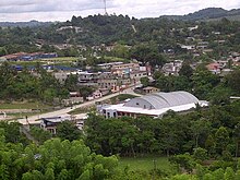 View across a wide, shallow valley with scattered buildings interspersed with dense patches of trees, and open, grassy spaces. The valley rises to a low, forested ridgeline. One of the hills supports a tall broadcasting mast.