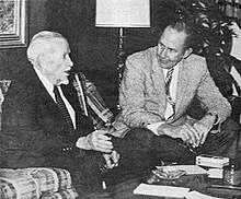 A black-and-white photograph of two men sitting by a low table talking to each other. The man on the left is much older, has white hair, and is wearing a dark suit with a white shirt and a dark tie. He sitting on a plaid couch and gesturing with his right hand as he speaks. The man on the left is younger, has dark hair, and is wearing a light jacket, dark pants, a white shirt, and a patterned tie. He is sitting on a chair with his arms resting on his legs as he leans forward to listen to the other man.