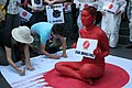 An oriental woman has painted herself red holding a sign (while sitting down) that says "Ban Whaling" while a crowd around her signs a petition. She is sitting on a Japanese flag with red dripping down (presumably to symbolize blood) (from Baleen whale)