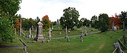 A panoramic view of the cemetery; a dirt road pass in front of the view from right to left. At center is a grey church-like structure, surrounded on all sides by various sized grave stones.
